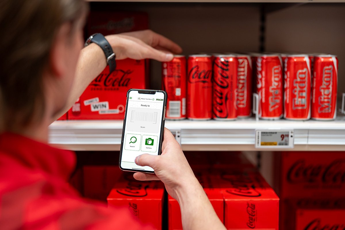 Retail worker using a smartphone app to scan a barcode on a grocery shelf with Coca-Cola cans for inventory management and digital pricing updates.