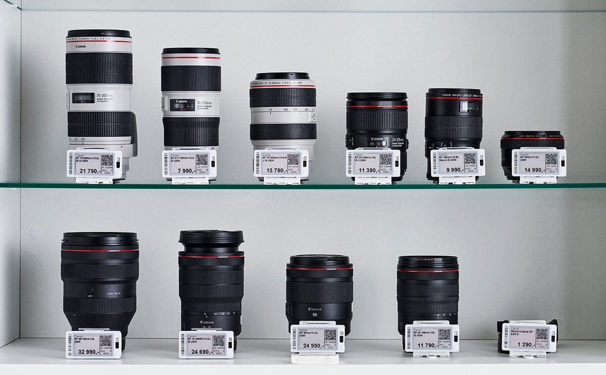 Professional Canon camera lenses, including EF and RF series, displayed on white shelves with electronic price tags and QR codes in a photography equipment store.